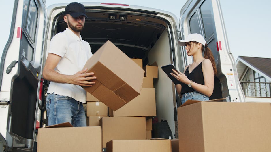 A man with a beard, wearing a pink cap, a two-tone beige and brown jacket over a checked shirt, and striped gloves, stands inside the open rear of a van during a home relocation. Behind him, the van's
