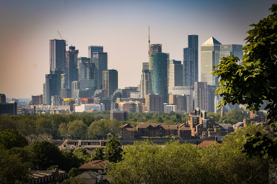 A wide view of a city skyline with numerous modern skyscrapers, some under construction with cranes visible at the top, towering over a lower area of residential buildings and lush green trees in the